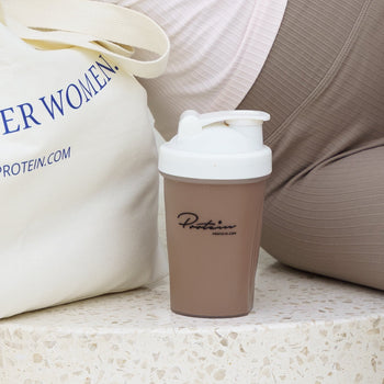 Brown protein shaker with white lid on a surface next to a beige bag with text.