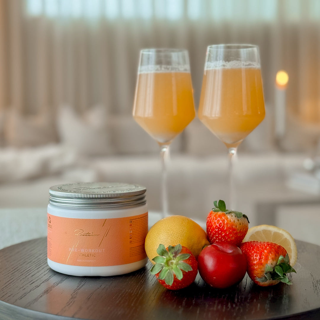 Two glasses of orange juice with a jar and fruits on a table in a blurred indoor setting
