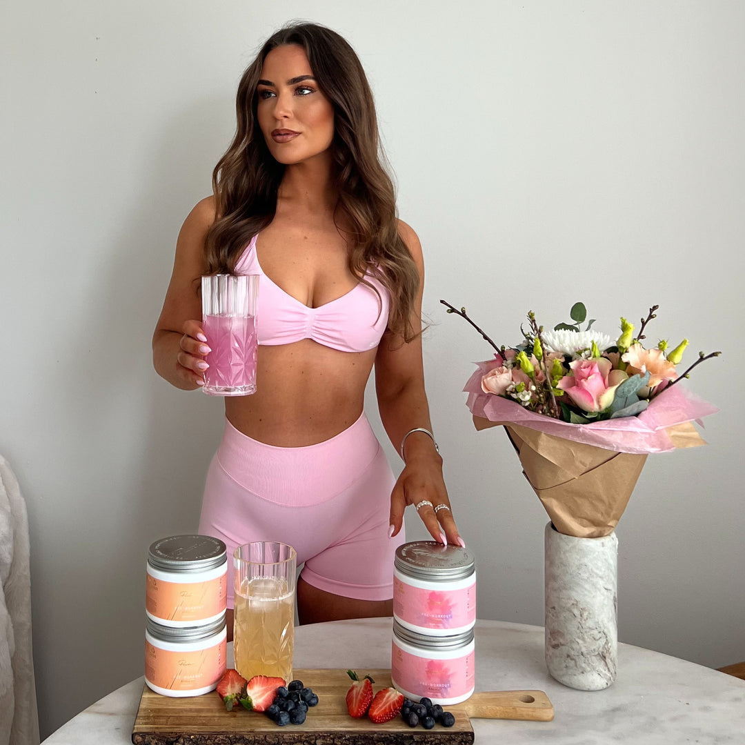 Woman in pink athletic wear holding a glass of pink drink with a bouquet of flowers and jars on a table.
